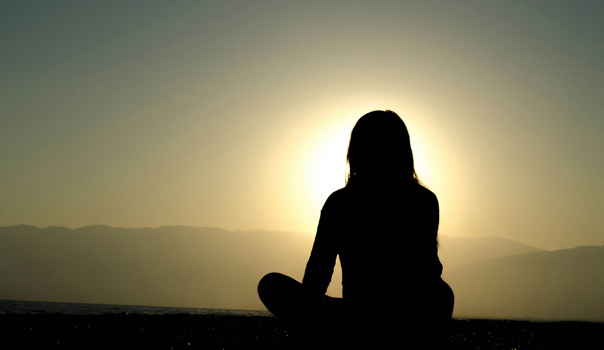 Woman sitting cross-legged in silhouette facing yellow sunset/sunrise
