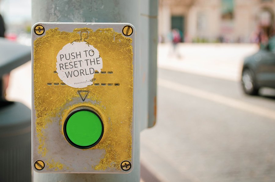 Well worn yellow crosswalk market with bright green button - sticker above says "Push to Reset the World." Photo: @joseantoniogall, Sticker: @Space_utopian